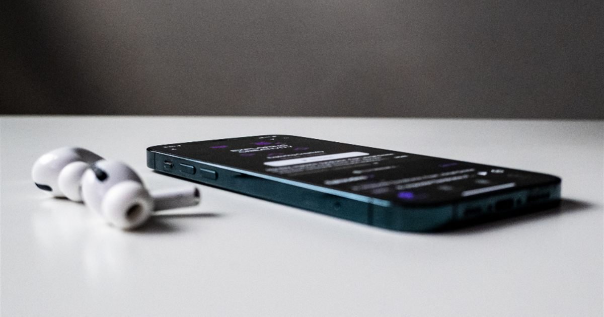 Celular deitado sobre uma mesa branca, ao lado de fones de ouvido sem fio