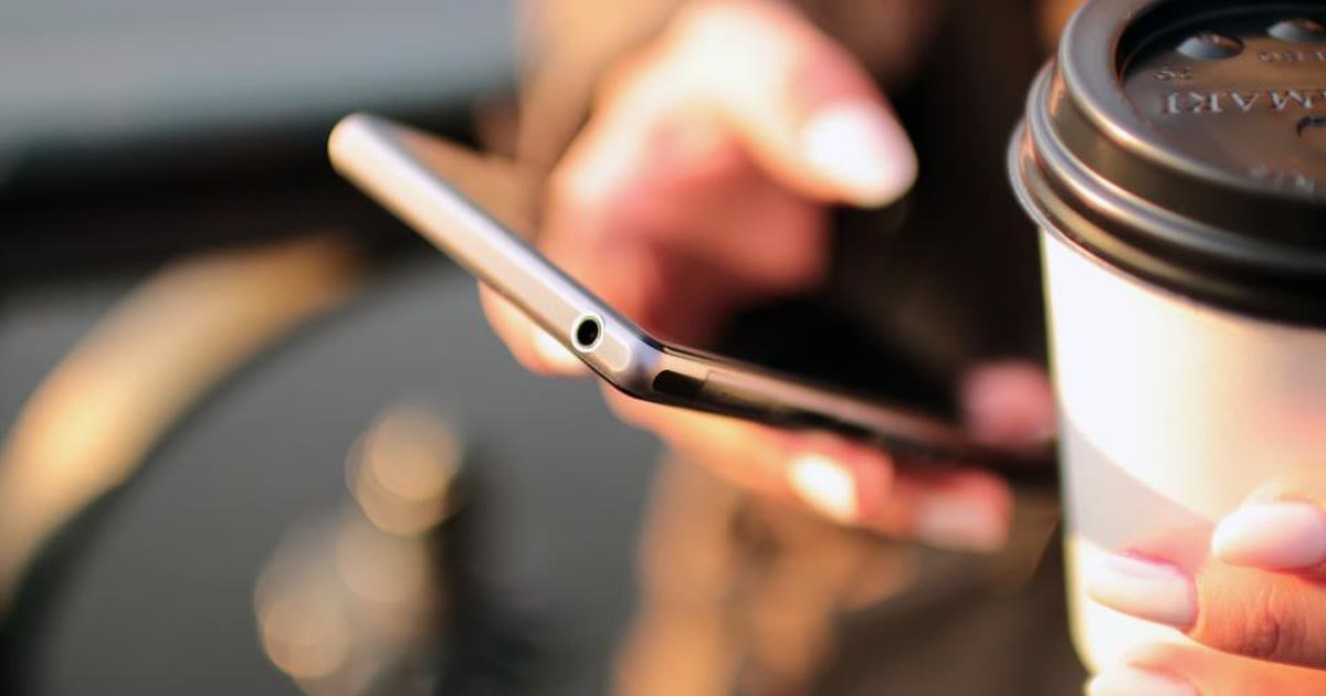 Mão segurando um celular, com um copo de café ao lado